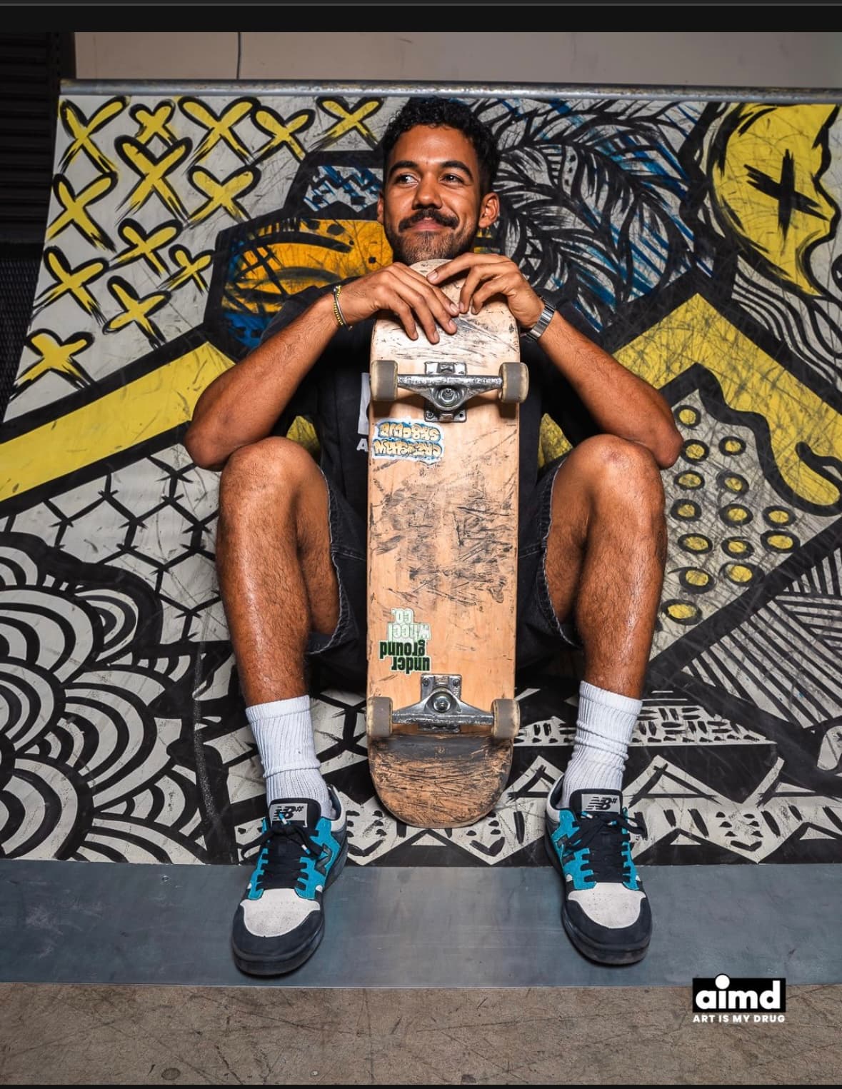 Smiling man with a mustache sits holding a skateboard against a black and yellow mural.